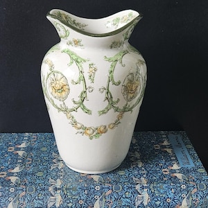 May include: A white ceramic vase with a flared rim and a decorative floral pattern in yellow and green. The vase has a slightly curved shape and a dark green trim around the top.