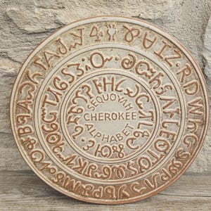 May include: A circular ceramic plaque featuring the Cherokee syllabary characters arranged in concentric rings. The center displays the text "SEQUOYAH CHEROKEE ALPHABET" in raised, brown lettering against a light, textured off-white background.