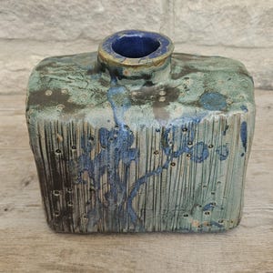 May include: A rectangular ceramic vase with a textured surface featuring vertical grooves and small indentations. The glaze is a mottled mix of sage green, dark brown, and deep blue, with blue drips running down the front and a solid blue interior.