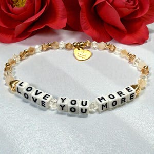 May include: A beaded bracelet with the words "LOVE YOU MORE" spelled out in white letter beads. The bracelet features gold-colored beads and a small gold heart charm. The background includes red roses.
