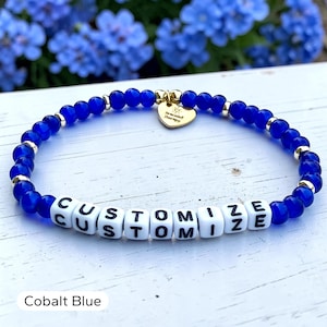 May include: A beaded bracelet featuring round cobalt blue beads, small gold-toned metallic spacers, and white square letter beads spelling out the word CUSTOMIZE. A gold-toned heart-shaped charm is attached, engraved with the text Bracelet Therapy.