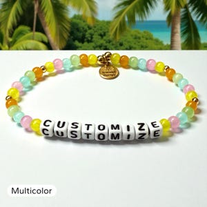 May include: A beaded bracelet featuring a repeating pattern of translucent yellow, orange, pink, and light green beads interspersed with small gold-tone spheres. A round gold-tone charm reads Bracelet Therapy. White square beads spell CUSTOMIZE.
