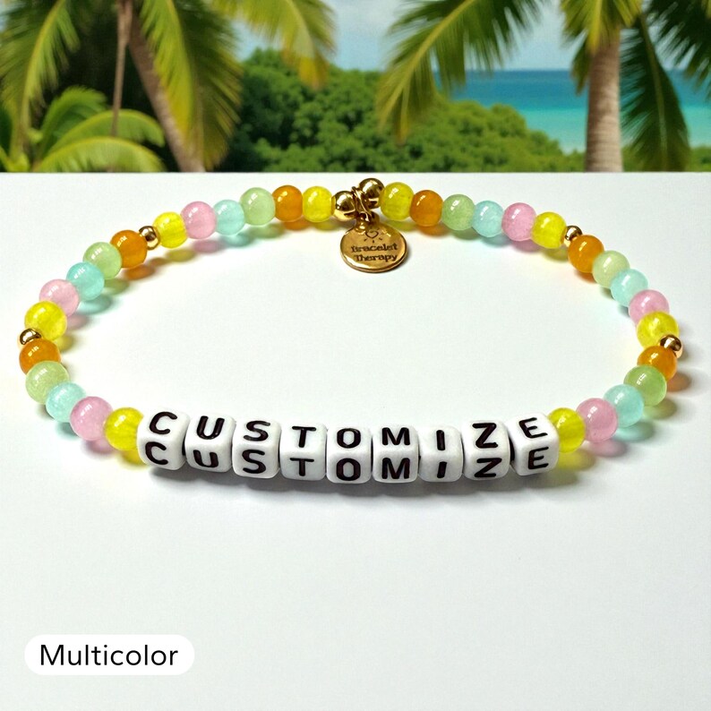 May include: A beaded bracelet featuring a repeating pattern of translucent yellow, orange, pink, and light green beads interspersed with small gold-tone spheres. A round gold-tone charm reads Bracelet Therapy. White square beads spell CUSTOMIZE.