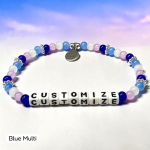 May include: A beaded bracelet featuring a repeating pattern of light blue, dark blue, white, and pale pink round beads interspersed with silver-toned spacers. The bracelet includes white square letter beads spelling CUSTOMIZE.