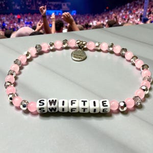 May include: A beaded bracelet featuring light pink round beads, faceted silver-toned beads, and white square letter beads spelling SWIFTIE. A round silver-toned metal charm hangs from the top, engraved with the text Bracelet Therapy.