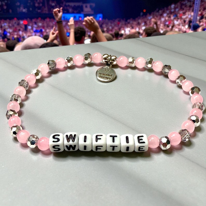 May include: A beaded bracelet featuring light pink round beads, faceted silver-toned beads, and white square letter beads spelling SWIFTIE. A round silver-toned metal charm hangs from the top, engraved with the text Bracelet Therapy.