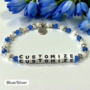 May include: A beaded bracelet featuring alternating blue, white, and silver-toned beads. The center displays white square beads with the black text CUSTOMIZE. A small round silver-toned metal charm hangs from the top, engraved with the text Bracelet Therapy.