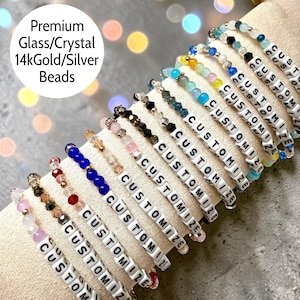 May include: A collection of beaded bracelets featuring white letter beads spelling CUSTOMIZE, interspersed with various colored glass and crystal beads, including blue, pink, yellow, and clear tones, accented with metallic gold and silver beads.