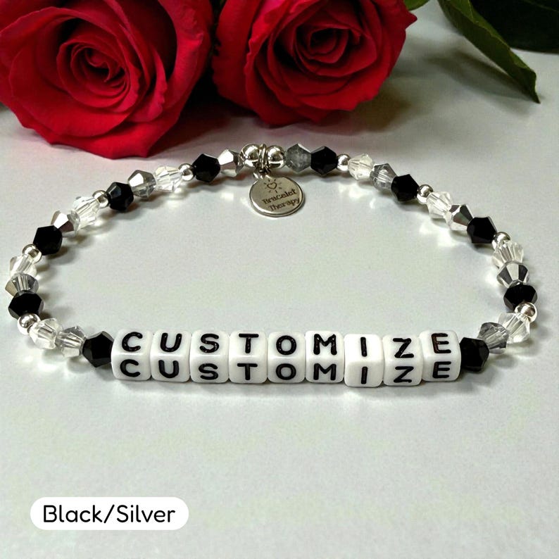 May include: A beaded bracelet featuring alternating black and clear faceted beads with silver-toned spacers. The center displays white square beads with the black text CUSTOMIZE. A small round silver charm reads Bracelet Therapy.
