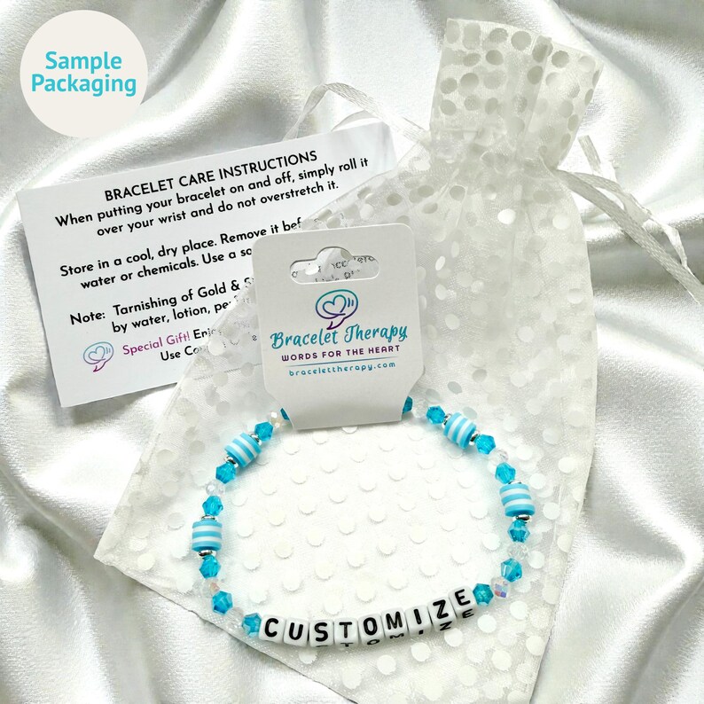 May include: A beaded bracelet featuring light blue and white striped cylindrical beads, clear faceted beads, and white letter beads spelling CUSTOMIZE. The bracelet is attached to a Bracelet Therapy display card next to a care instruction card.