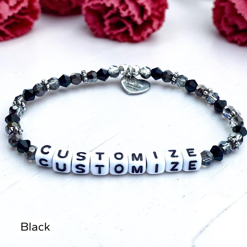 May include: A beaded bracelet featuring alternating black and dark gray faceted crystal beads, silver-toned spacers, and white square letter beads spelling the word CUSTOMIZE. A small silver heart-shaped charm hangs from the top of the bracelet.