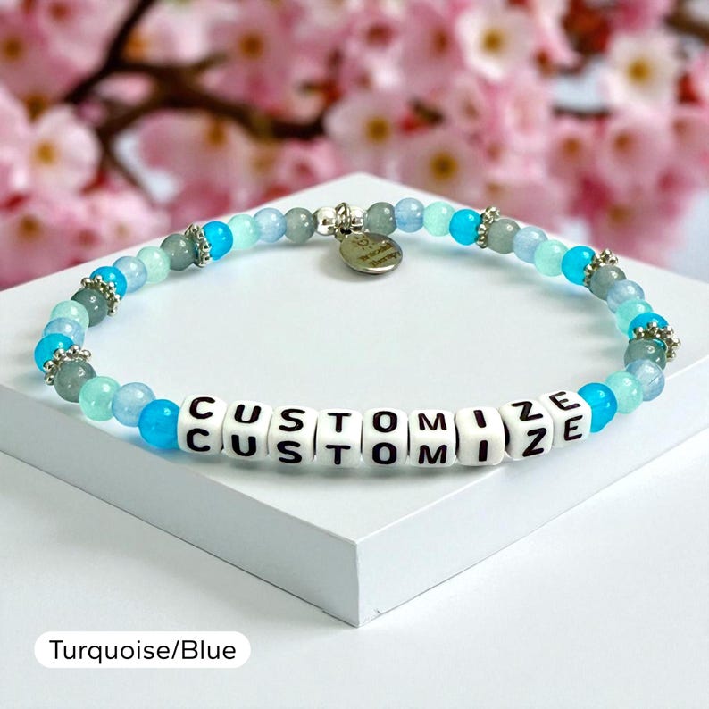 May include: A beaded bracelet featuring alternating shades of blue and turquoise round beads, interspersed with small silver-toned metal spacers. The center includes white square beads with black letters spelling out the word CUSTOMIZE.