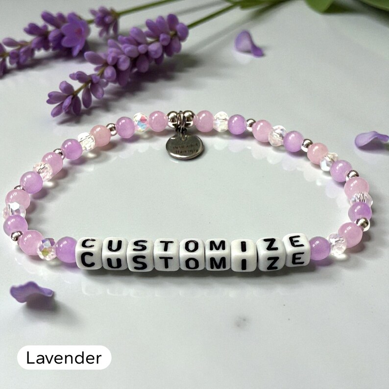 May include: A beaded bracelet featuring light purple and clear faceted beads, interspersed with small silver-toned spacers. The center displays white square beads with the black text CUSTOMIZE, and a small round silver-toned metal charm is attached.