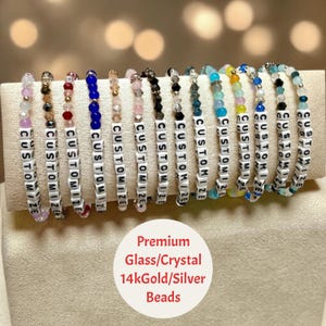May include: A collection of thirteen beaded bracelets featuring colorful glass and crystal beads, metallic accents, and white square letter beads spelling the word CUSTOMIZE. The bracelets are displayed in a row on a beige fabric stand.