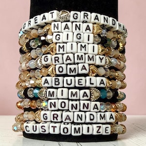 May include: Stack of beaded bracelets with white letter beads spelling out "GREAT GRANDMA", "NANA", "GIGI", "MIMI", "GRAMMY", "OMA", "ABUELA", "MIMA", "NONNA", "GRANDMA", and "CUSTOMIZE". The bracelets feature gold and crystal accents.