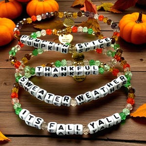 May include: Four elastic beaded bracelets featuring translucent orange, green, and brown glass beads. Each bracelet includes white square letter beads spelling PUMPKIN SPICE, THANKFUL, SWEATER WEATHER, and ITS FALL YALL, with small gold heart charms.