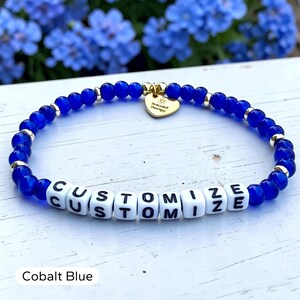 May include: A beaded bracelet featuring round cobalt blue beads, small gold-toned metallic spacers, and white square letter beads spelling out the word CUSTOMIZE. A gold-toned heart-shaped charm is attached, engraved with the text Bracelet Therapy.