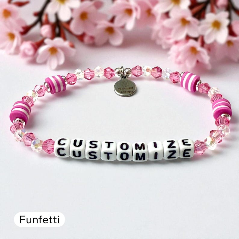 May include: A beaded bracelet featuring pink and white striped cylindrical beads, translucent light pink faceted crystal beads, and white square letter beads spelling out the word CUSTOMIZE. A small round silver metal charm is attached to the top.