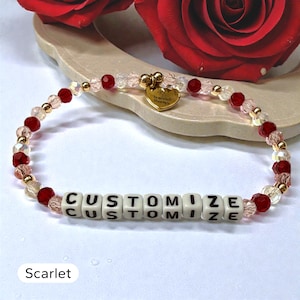 May include: A beaded bracelet featuring alternating red and clear faceted beads, small gold-toned spacer beads, and white square letter beads spelling out the word CUSTOMIZE. A small gold-toned heart-shaped charm hangs from the top of the bracelet.