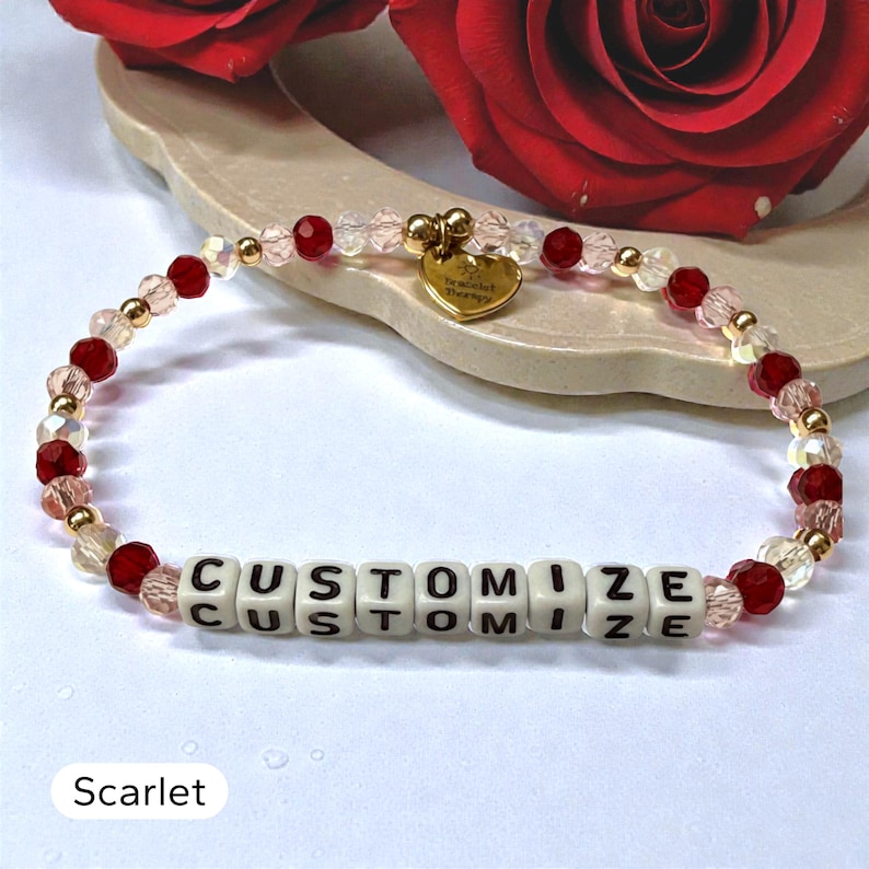 May include: A beaded bracelet featuring alternating red and clear faceted beads, small gold-toned spacer beads, and white square letter beads spelling out the word CUSTOMIZE. A small gold-toned heart-shaped charm hangs from the top of the bracelet.
