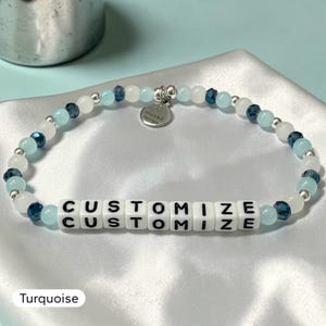 May include: A beaded bracelet featuring alternating light blue, white, and dark blue faceted beads with small silver spacers. A central section contains white square beads with the black text CUSTOMIZE. A silver circular charm reads bracelet therapy.