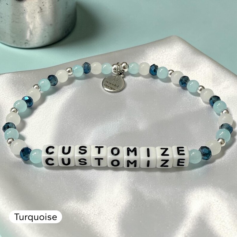May include: A beaded bracelet featuring alternating light blue, white, and dark blue faceted beads with small silver spacers. A central section contains white square beads with the black text CUSTOMIZE. A silver circular charm reads bracelet therapy.
