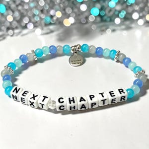 May include: A beaded bracelet featuring alternating light blue, dark blue, and translucent gray beads with silver-toned spacers. The bracelet includes white square letter beads spelling NEXT CHAPTER and a round silver charm engraved with Bracelet Therapy.