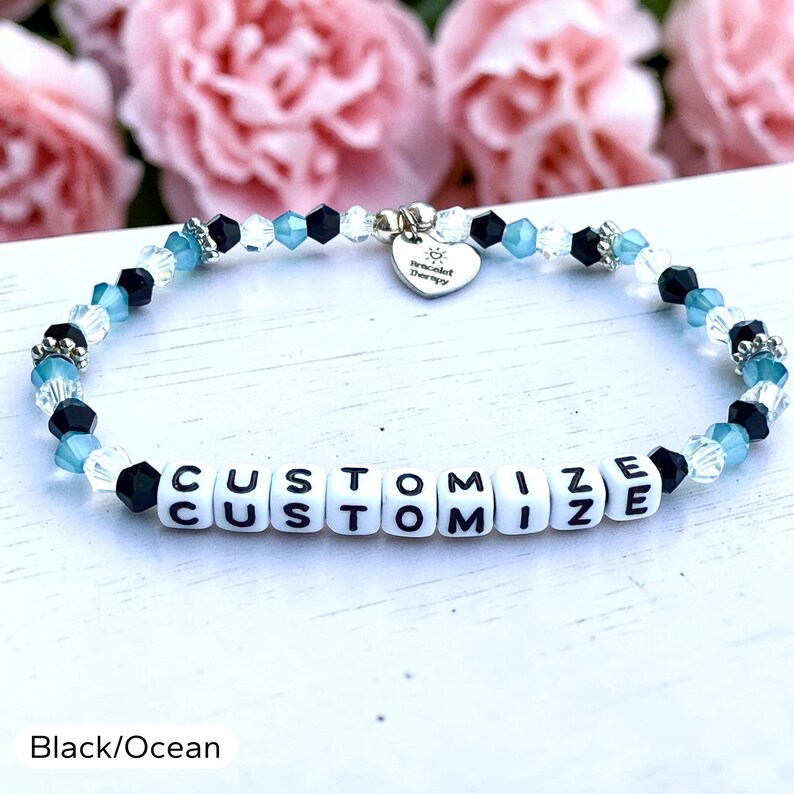 May include: A beaded bracelet featuring alternating black, clear, and ocean blue faceted beads. The bracelet includes white square letter beads spelling out the word CUSTOMIZE and a silver heart-shaped charm engraved with the text Bracelet Therapy.