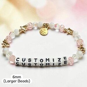 May include: A beaded bracelet featuring white and light pink faceted beads, gold-toned star charms, and white square beads with the black text CUSTOMIZE. A round gold-toned charm reads Bracelet Therapy. The beads measure 6mm in diameter.