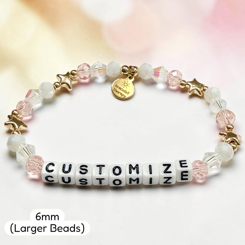May include: A beaded bracelet featuring white and light pink faceted beads, gold-toned star charms, and white square beads with the black text CUSTOMIZE. A round gold-toned charm reads Bracelet Therapy. The beads measure 6mm in diameter.