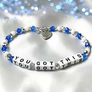 May include: A beaded bracelet featuring white and blue beads, silver-toned accents, and white square letter beads spelling out YOU GOT THIS. A silver heart-shaped charm hangs from the center, engraved with the text Bracelet Therapy.