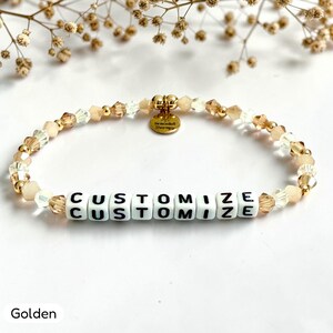 May include: A beaded bracelet featuring alternating clear and champagne-colored faceted crystals, small gold-toned metallic beads, and white square letter beads spelling CUSTOMIZE. A small round gold-toned metal charm hangs from the center.