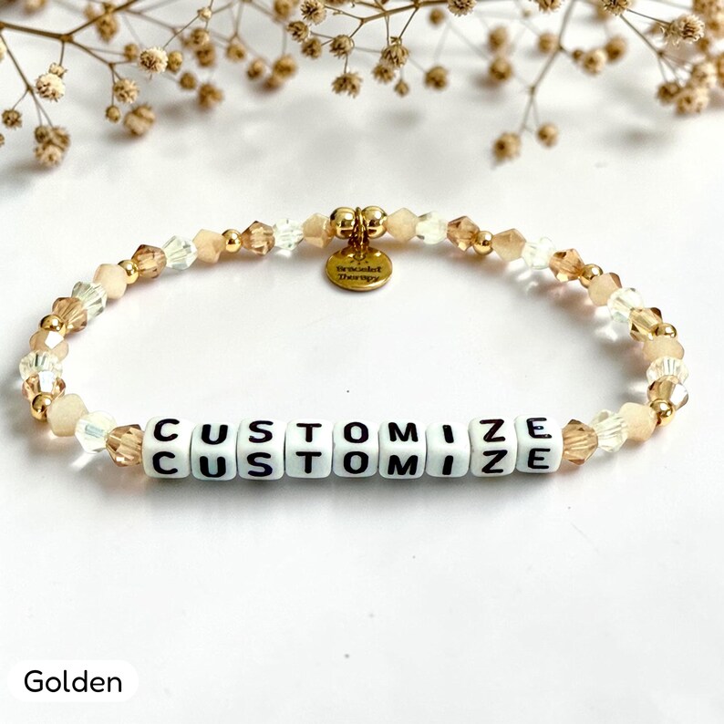 May include: A beaded bracelet featuring alternating clear and champagne-colored faceted crystals, small gold-toned metallic beads, and white square letter beads spelling CUSTOMIZE. A small round gold-toned metal charm hangs from the center.