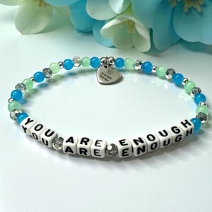May include: A beaded bracelet featuring alternating light blue, bright blue, and clear faceted beads. White square letter beads spell out the phrase YOU ARE ENOUGH. A silver heart-shaped charm is attached, engraved with the text Bracelet Therapy.