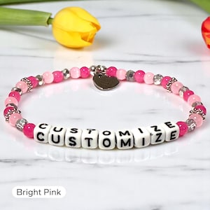 May include: A beaded bracelet featuring alternating bright pink and translucent beads, silver-toned spacers, and a central heart-shaped charm. White square beads with black lettering spell out the word CUSTOMIZE in the center of the bracelet.