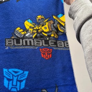 May include: Blue fleece blanket featuring the Transformers character Bumblebee. The blanket has a repeating pattern of Bumblebee, Autobot symbols, and the word "BUMBLEBEE" in gray lettering. The blanket is held up by a person.