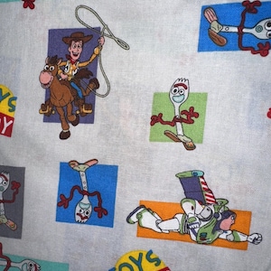 K&ouml;nnte beinhalten: Wei&szlig;er Stoff mit bunten Cartoon-Charakteren aus dem Film Toy Story. Das Design umfasst Woody auf einem Pferd, Forky und Buzz Lightyear. Die Worte "TOYS at PLAY" sind ebenfalls sichtbar.