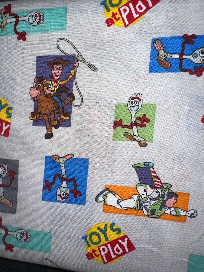 K&ouml;nnte beinhalten: Wei&szlig;er Stoff mit bunten Cartoon-Charakteren aus dem Film Toy Story. Das Design umfasst Woody auf einem Pferd, Forky und Buzz Lightyear. Die Worte "TOYS at PLAY" sind ebenfalls sichtbar.