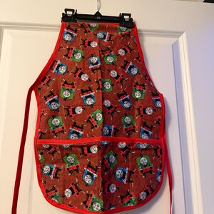 May include: A red apron with a repeating pattern of cartoon train faces. The apron has a red border and red ties. Two pockets are visible on the front of the apron. The apron is hanging on a black hanger.