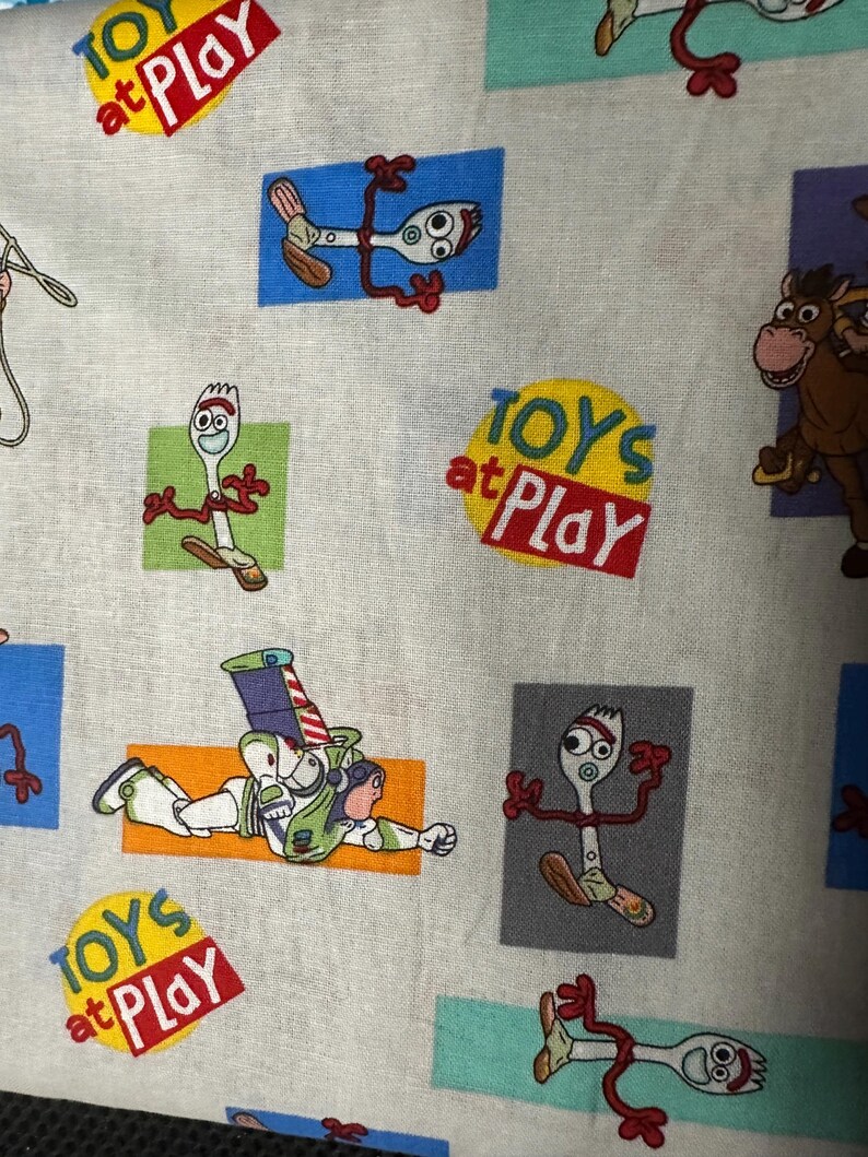 K&ouml;nnte beinhalten: Stoff mit einem sich wiederholenden Muster von Charakteren aus dem Film Toy Story. Das Design umfasst Bilder von Forky, Buzz Lightyear und einem Pferd sowie den Text "TOYS at PLAY" in einem roten und gelben Kreis.