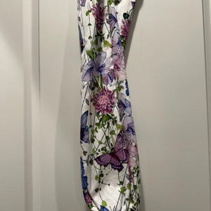 Puede incluir: Una bolsa de tela blanca con un estampado floral y de mariposas. El dise&ntilde;o presenta flores y mariposas moradas, rosas y azules sobre un fondo blanco. La bolsa tiene un fondo fruncido.