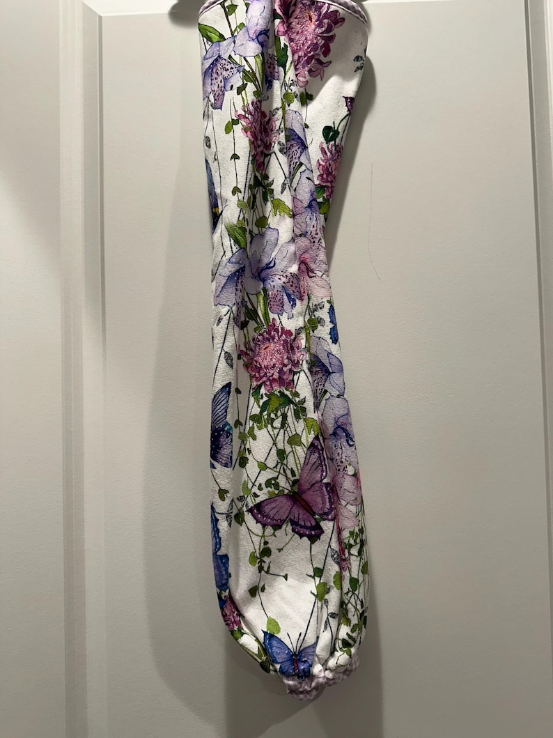 Puede incluir: Una bolsa de tela blanca con un estampado floral y de mariposas. El dise&ntilde;o presenta flores y mariposas moradas, rosas y azules sobre un fondo blanco. La bolsa tiene un fondo fruncido.