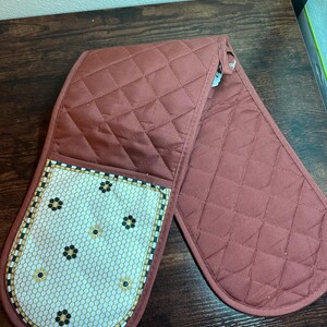 May include: A quilted, brick-red double oven mitt. One side features a white and beige mosaic pattern with black and gold floral accents. The mitt is designed with a diamond pattern and a loop for hanging.