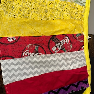 May include: A patchwork apron with a variety of fabrics. The apron features yellow paisley, red Coca-Cola print, gray chevron, and solid red and purple fabrics. A yellow border and ties complete the design.