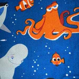 May include: Blue fabric featuring cartoon sea creatures. An orange octopus, clownfish, and other fish swim among white bubbles. A white beluga whale and a black and yellow fish are also present.