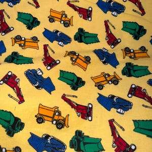 May include: Yellow fabric with a repeating pattern of colorful construction vehicles, including dump trucks, excavators, and bulldozers in red, blue, green, and yellow. The vehicles are arranged in various orientations.