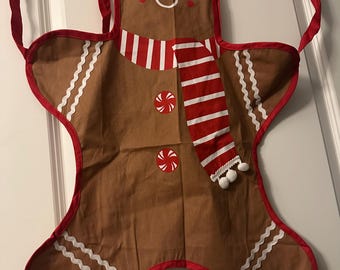 May include: A brown gingerbread man apron with a red and white striped scarf and red trim. The apron has a smiling face, red cheeks, and white icing details. Two peppermint candies are on the apron.