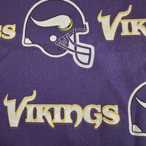 May include: Purple fabric featuring a repeating pattern of white and gold football helmets and the word "Vikings" in a stylized font. The design is a repeating pattern across the fabric.