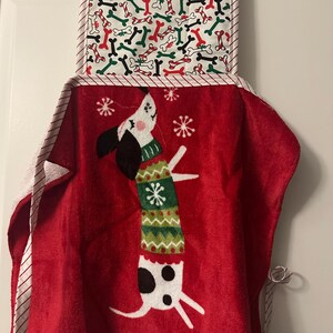 May include: A red kitchen towel with a dachshund dog wearing a green and white sweater, and snowflakes. The top section has a pattern of bones in red, green, and white. The towel is hanging on a black bar.