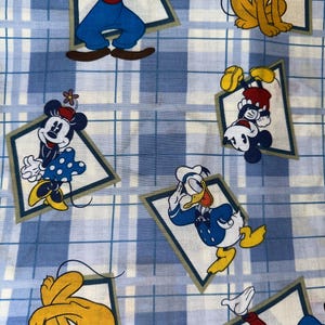 May include: Fabric featuring cartoon characters including Mickey Mouse, Minnie Mouse, Donald Duck, Goofy, and Pluto. The characters are set within diamond-shaped frames against a blue and white plaid background.